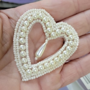 以下が含まれることがあります： 白いシードビーズと模造真珠で装飾された、涙の形をした真珠が付いたハート型のアップリケ。 このクラフトアイテムは、おそらく衣類やクラフトプロジェクト用です。