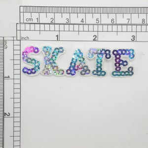 May include: An iron-on patch featuring the word "SKATE" in iridescent blue, purple and pink sequins.