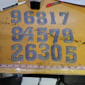 May include: A collection of denim-style fabric numbers, including 9, 6, 8, 1, 7, 8, 4, 5, 7, 9, 2, 6, 3, 0, and 5. The numbers are blue with yellow stitching, arranged on a wooden surface. A ruler and pens are also visible.