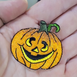 Può includere: Una toppa ricamata a forma di zucca di Halloween. La zucca è di colore arancione brillante con un volto sorridente giallo, occhi triangolari e una bocca ampia e curva. Presenta un gambo verde scuro con un piccolo dettaglio a viticcio.