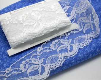 May include: Two white lace trims on a blue fabric background. The top trim is a delicate floral pattern and the bottom trim is a wider, more elaborate floral pattern. The text "Patchwork Panda Trims" is visible on the white card.