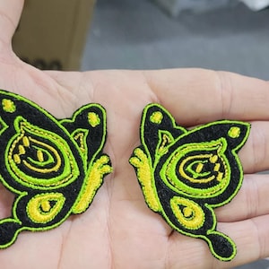 May include: Two embroidered butterfly patches featuring a black base with vibrant neon green and yellow thread detailing. The butterfly wings display intricate circular and curved patterns outlined in bright green, creating a high-contrast design.