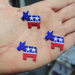 May include: Three embroidered donkey patches in red, white, and blue. The donkeys have blue bodies with white stars and red legs. These patches are likely for political or patriotic themed projects.