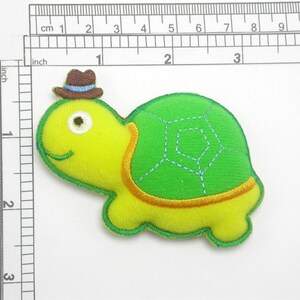 May include: A green and yellow felt turtle applique with a brown felt cowboy hat. The turtle has a stitched-on smile and a green shell with a geometric pattern.