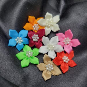 Peut inclure: Assortiment de neuf fleurs en ruban de satin avec des centres de perles. Les fleurs sont de différentes couleurs, notamment bleu, vert, rouge, orange, blanc et rose.