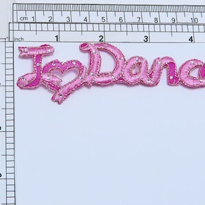 Può includere: Applique in tessuto rosa con le parole "I Love Dance" in un carattere corsivo con strass.