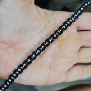 Op de afbeelding: Een close-up van een zwart koordarmband met zilverkleurige kralen. De armband is gemaakt van een geweven zwart koord met gelijkmatig verdeelde, kleine, ronde zilveren kralen. De armband wordt in een hand gehouden.