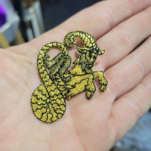 Pode incluir: Patch bordado em ouro e preto com o símbolo do zodíaco Capricórnio. O design retrata uma criatura mítica com cabeça de cabra e cauda de peixe. O patch é detalhado com costuras intrincadas.