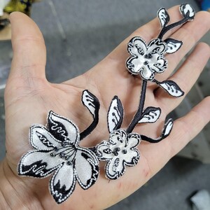 May include: A floral applique with white satin flowers and black embroidered details. The applique features three flowers and several leaves on a black stem. Small beads are in the center of each flower.