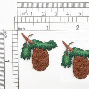 Pode incluir: Dois apliques bordados de pinhas marrons e verdes com um galho e folhas. As pinhas têm um diâmetro de aproximadamente 2,5 cm.