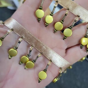 May include: Close-up of decorative trim featuring a tan ribbon with dangling yellow disc-shaped beads. The beads are accented with small, clear crystal beads and antique brass-colored metal details. This trim is suitable for various craft and sewing projects.