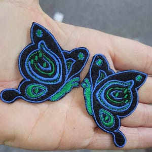 May include: Two embroidered butterfly patches featuring a black felt base with intricate stitching in vibrant blue and green thread. The butterfly designs display detailed wing patterns with concentric shapes and small circular accents in bright green.