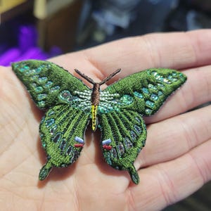 May include: Embroidered green butterfly patch with detailed wings and a brown body. The wings feature iridescent sequins and intricate stitching. The butterfly has a yellow stripe down the center of its body.