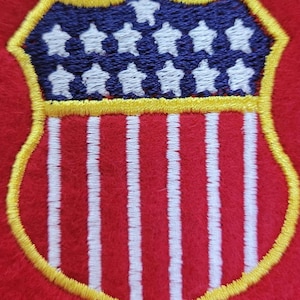 May include: A shield-shaped embroidered patch on a red felt background. The patch features a blue upper section with white embroidered stars and a lower section with vertical white stripes, all outlined in a bright yellow thread border.