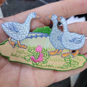 May include: Embroidered patch featuring three light blue geese with orange beaks and feet, standing on a beige and green landscape with pink flowers. The patch has a detailed, textured design, suitable for fabric embellishment.