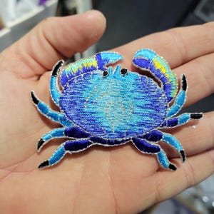 Op de afbeelding: Geborduurde krabpatch in blauwtinten, met gele accenten op de scharen en zwarte punten. De krab heeft een gedetailleerd ontwerp, perfect om toe te voegen aan kleding of accessoires. De patch is ongeveer 7.6 cm breed.