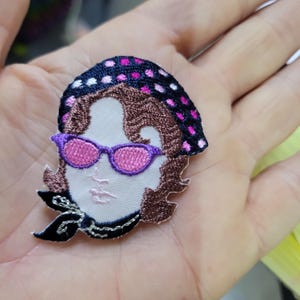 Op de afbeelding: Geborduurde patch met een gestileerd gezicht met paarse zonnebril, een zwart en roze gestippelde hoofddoek en bruin haar. De patch heeft een wit gezicht en een zwarte sjaal. Geschikt voor kleding of accessoires.