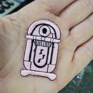 Könnte beinhalten: Ein rosa gestickter Aufnäher in Form eines Retro-Jukebox. Der Aufnäher hat schwarze Details, darunter der Lautsprechergrill, Knöpfe und Noten. Der Aufnäher ist wahrscheinlich für Kleidung oder Accessoires.