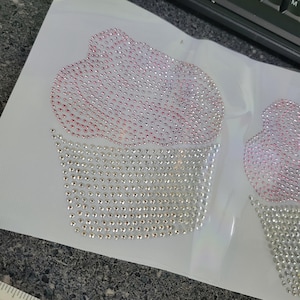 Puede incluir: Dos parches termoadhesivos de cupcake con pedrería. La cobertura del cupcake es rosa y el envoltorio es plateado. Los brillantes están dispuestos para crear la forma de un cupcake. Los parches están sobre un respaldo blanco.