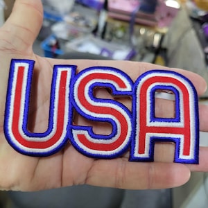 May include: An embroidered patch featuring the letters USA in a bold, block-style font. The letters are colored in bright red with a thick white outline and a dark blue border, creating a layered, patriotic appearance suitable for fabric application.