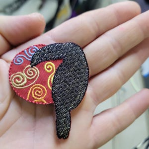 May include: A decorative embroidered patch featuring a black bird silhouette with a red and gold circular design. The patch has a glittery texture and is likely designed for clothing or accessories.