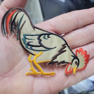 May include: Embroidered patch of a rooster in beige, brown, and teal. The rooster has a red comb and tail feathers with orange accents. The patch features yellow legs and a curved posture, suitable for clothing or crafts.