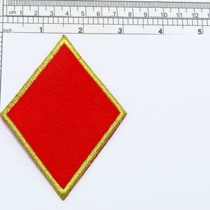 Könnte beinhalten: Ein roter, rautenförmiger Patch mit goldener Umrandung. Der Patch ist etwa 5 cm breit.