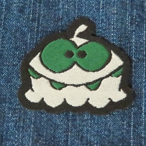 May include: White and green embroidered patch of a cartoon monster with a toothy grin. The monster has large green eyes and a black outline.