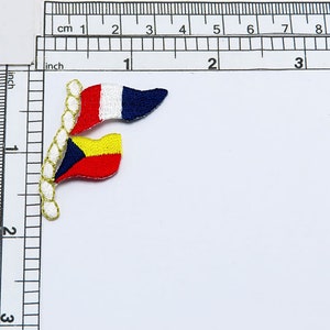 Op de afbeelding: Twee kleine stoffen vlaggen aan elkaar genaaid met een gouden touw. De bovenste vlag is blauw, wit en rood. De onderste vlag is rood, geel en blauw.