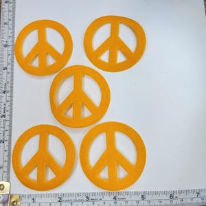Peut inclure: Cinq découpes de signes de paix orange. Les symboles de la paix sont faits d'un matériau semblable à du feutre. L'image comprend une règle avec des mesures en centimètres.