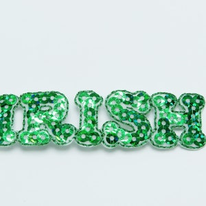 Könnte beinhalten: Das Wort "IRISH" in grünen Paillettenbuchstaben. Jeder Buchstabe ist weiß umrandet und mit glänzenden grünen Pailletten gefüllt. Die Buchstaben stehen vor einem weißen Hintergrund.
