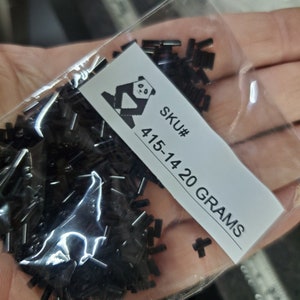 Puede incluir: Una bolsa de plástico transparente llena de pequeñas cuentas cilíndricas negras. Una etiqueta blanca con un logotipo de panda y el texto "SKU# 415-14 20 GRAMOS" es visible. Las cuentas están dispersas en la bolsa.