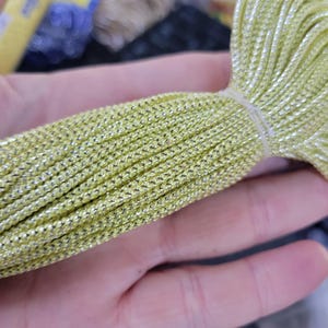 Può includere: Primo piano di un fascio di cordoncino metallico verde chiaro e argento, saldamente legato con un laccio bianco. Il cordoncino ha un aspetto intrecciato ed è tenuto in mano. Materiale per artigianato.