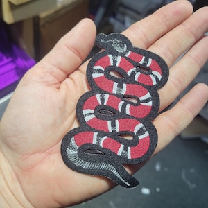 May include: A fabric patch in the shape of a coiled snake. The patch features a red body with white stripes, outlined in black. The snake's head is detailed with a gray and white pattern.