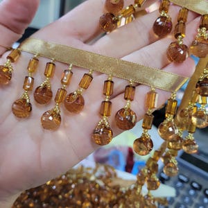 May include: Gold ribbon trim with dangling amber-colored beads. The trim features a satin ribbon base with alternating rectangular and spherical beads. The beads are faceted, catching light and adding a decorative element to the trim.