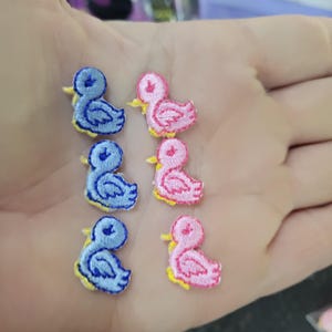 Może przedstawiać: Haftowane naszywki z kaczkami w kolorze niebieskim i różowym. Każda naszywka przedstawia wzór kaczki z żółtymi dziobami i stopami. Naszywki są małe, idealne do ozdabiania odzieży lub rękodzieła. Kaczki ułożone są we wzór.