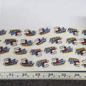 Op de afbeelding: Een vel wit papier met 30 kleine geborduurde pleisters van blauwe en rode sleepboten met gele accenten. De pleisters zijn in rijen gerangschikt en ongeveer 2,5 cm groot. Een liniaal is zichtbaar aan de onderkant van de afbeelding.