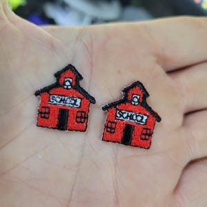 Op de afbeelding: Twee geborduurde patches in de vorm van rode schoolgebouwen met zwarte randen. Elke patch heeft het woord "SCHOOL" in witte letters. De patches hebben een klein raam en deuren.