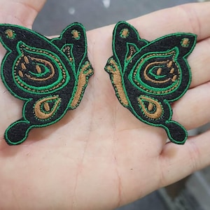 May include: Two embroidered butterfly patches featuring a black base with intricate green and bronze thread detailing. The wings display a symmetrical pattern with curved lines and circular accents, finished with a vibrant green stitched border.