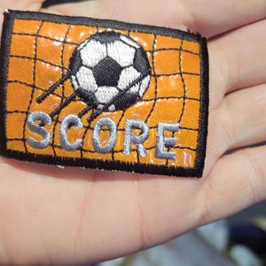 May include: A rectangular embroidered patch featuring an orange background with a black grid pattern representing a soccer goal net. A black and white soccer ball is depicted in the center, with the word SCORE stitched in silver thread below.