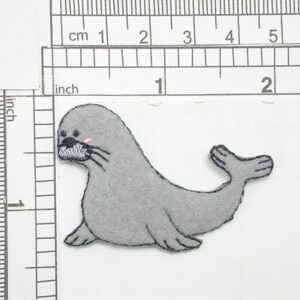 Può includere: Un'applicazione di foca in feltro grigio con un blush rosa sulla guancia. La foca è lunga circa 5 cm.
