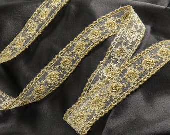 May include: Gold glitter trim lace ribbon with a delicate floral pattern. The ribbon is on a black fabric background.