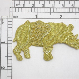 May include: A gold embroidered iron-on patch of a rhinoceros. The rhinoceros is facing right and has a detailed design.