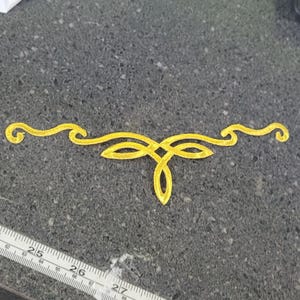 May include: A bright yellow embroidered appliqué featuring a symmetrical, flowing, knot-like design with curved ends. A white measuring tape is positioned below the appliqué, showing the design spans approximately 15 cm in total width.