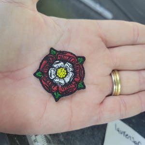 May include: Embroidered patch featuring a Tudor rose design. The patch has a red outer ring, white petals, and a yellow centre. Green leaves are at each corner, and the patch is outlined in black.