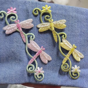 Op de afbeelding: Twee geborduurde strijkpatches met een libellenmotief. De patches bevinden zich op een blauwe denim stoffen achtergrond. Eén patch is roze en groen, de andere is geel en groen.