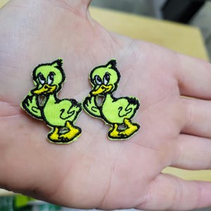 May include: Two embroidered patches featuring a cartoon duck design. The ducks are light green with black outlines and yellow beaks and feet. The patches are likely for clothing or accessories.