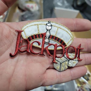 May include: Embroidered patch with the word "Jackpot" in red script. The patch features a roulette wheel design with a silver and white casino chip, and a pair of dice. The patch has gold and black accents.