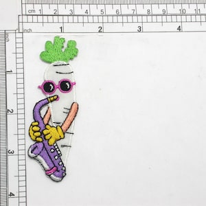 Pode incluir: Um remendo bordado de um rabanete branco com óculos de sol rosa tocando um saxofone roxo. O rabanete tem uma parte superior verde com folhas.