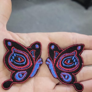 May include: Two embroidered butterfly patches featuring a black base with vibrant red and blue thread detailing. Each patch displays a symmetrical wing pattern with intricate stitched lines in red and blue, creating a distinct contrast against the black fabric background.
